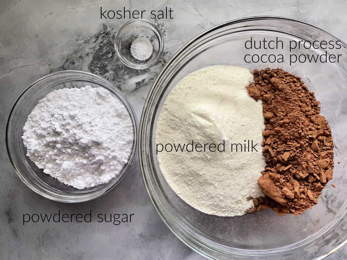 Ingredients on marble countertop: powder milk, cocoa powder, salt, and powder sugar.