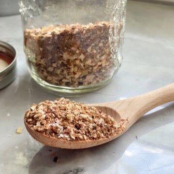 Glass jar in background with seasoning and wooden spoon with seasoning on marble countertop in foreground.