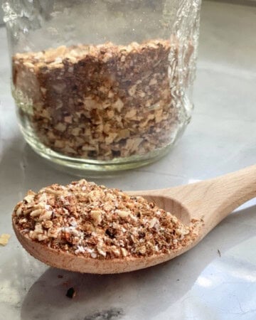 Glass jar in background with seasoning and wooden spoon with seasoning on marble countertop in foreground.
