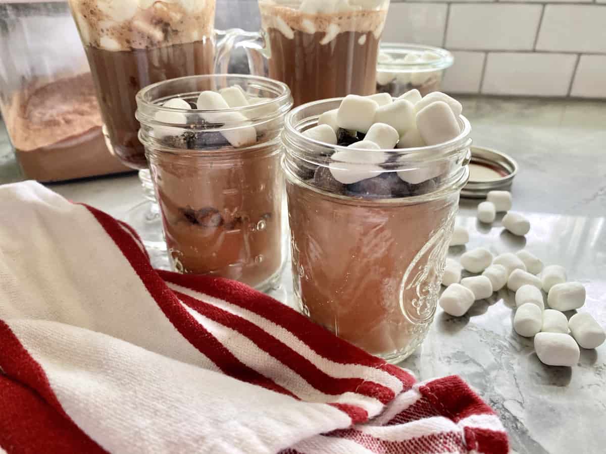 Two mason jars filled with hot chocolate powder mix with a red and white striped kitchen towel in the foreground.