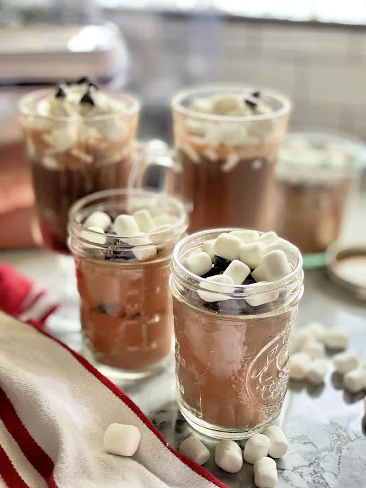 Two mason jars filled with cocoa mix and two glasses blurred out of hot chocolate in the background.