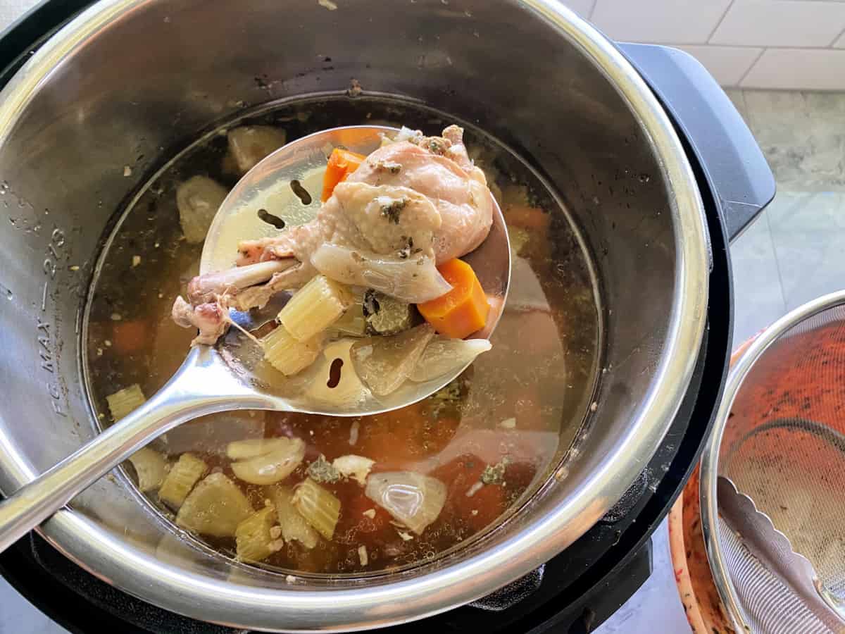 Slotted spoon holding cooked carrots, celery, onions, and a chicken drumstick over an Instant Pot.