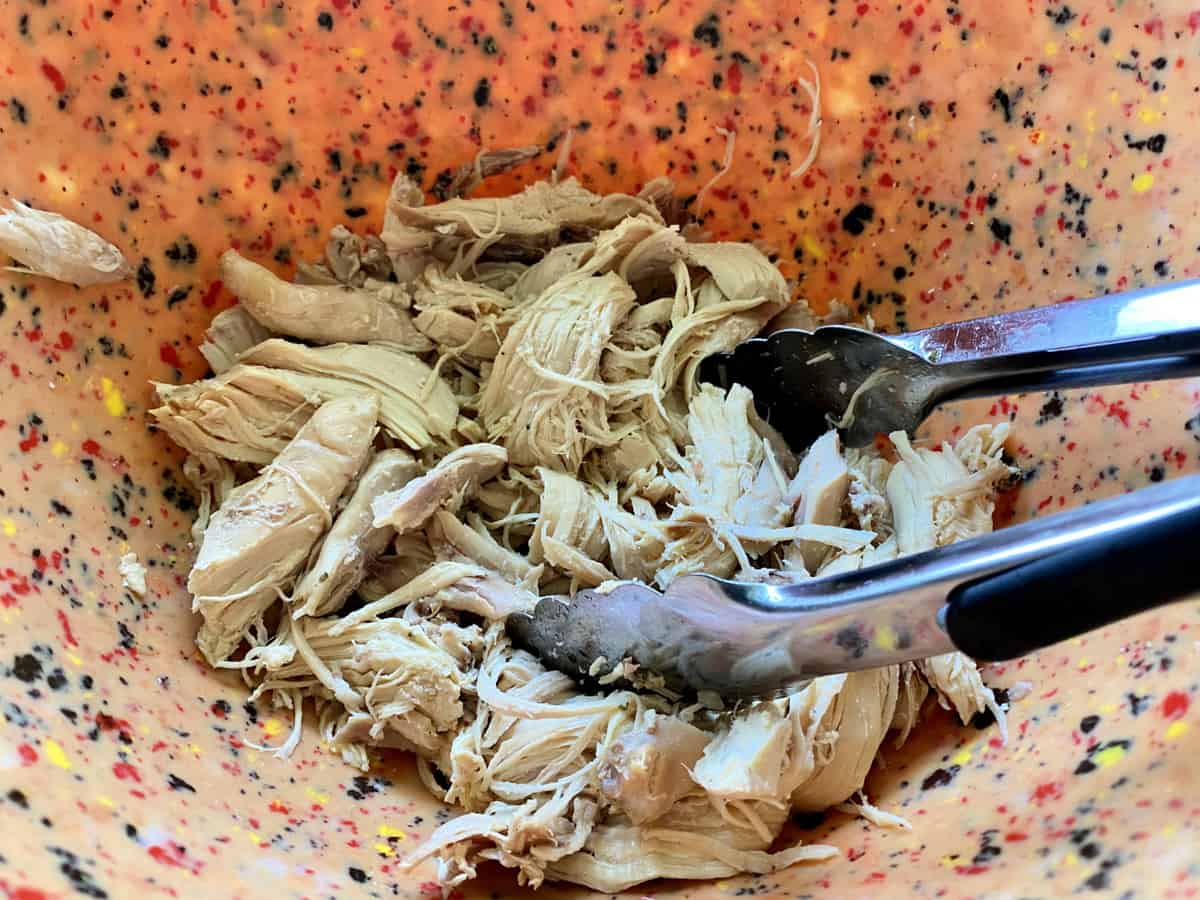 Tongs holding shredded chicken in an orange speckled bowl.