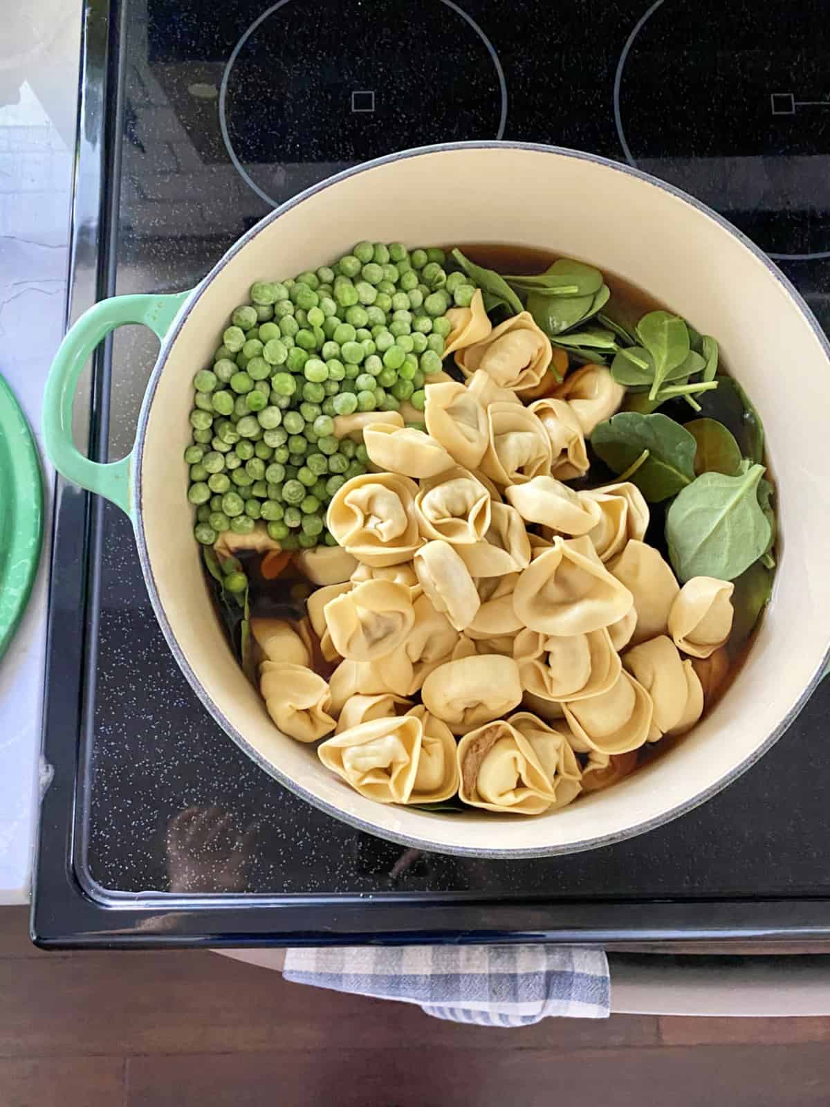 Top view of a green pot on a stove filled with tortellini, frozen peas, spinach leaves, and broth.