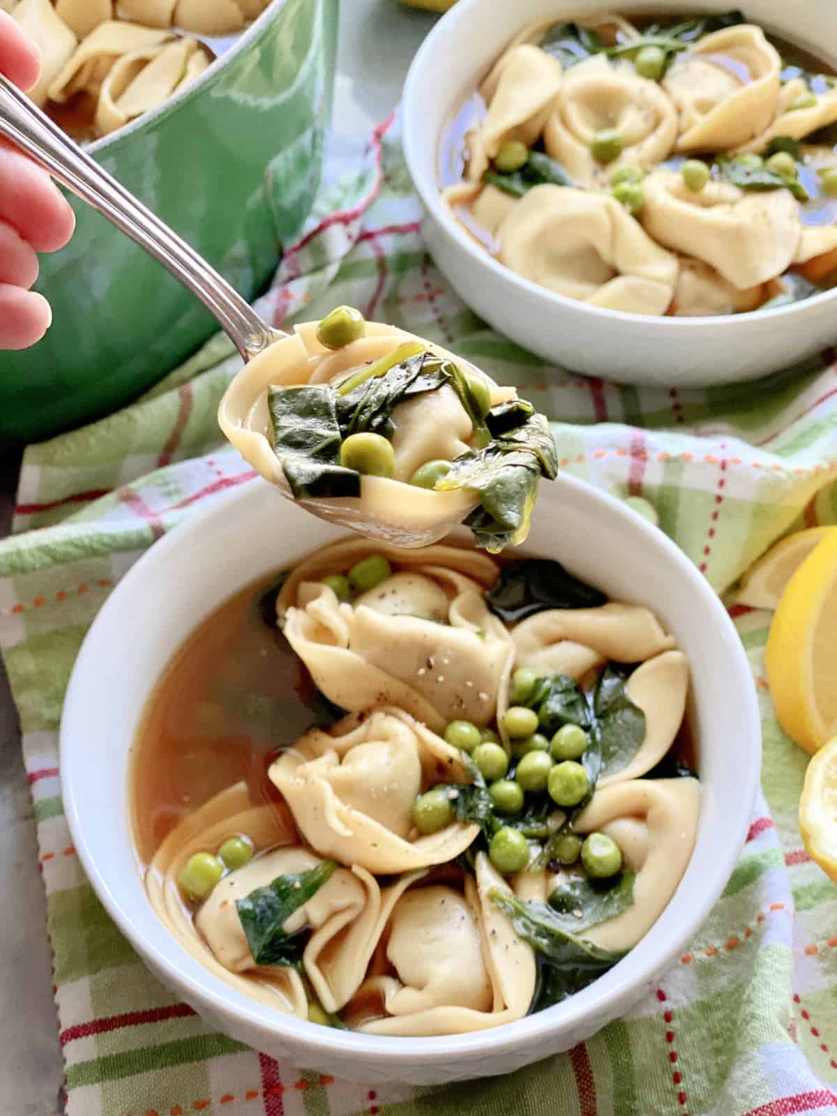 Female hand holding a spoonful of tortelini, spinach, and green peas over a bowl of soup.