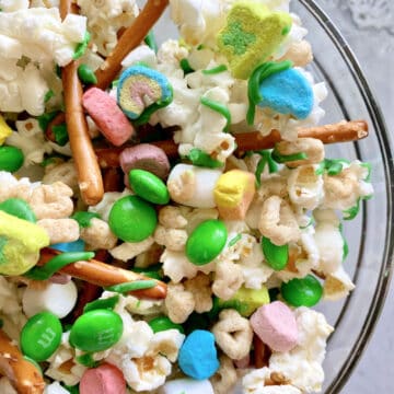 Close up of a glass bowl of popcorn, green M&M's, marshmallows, lucky charms, and pretzels.