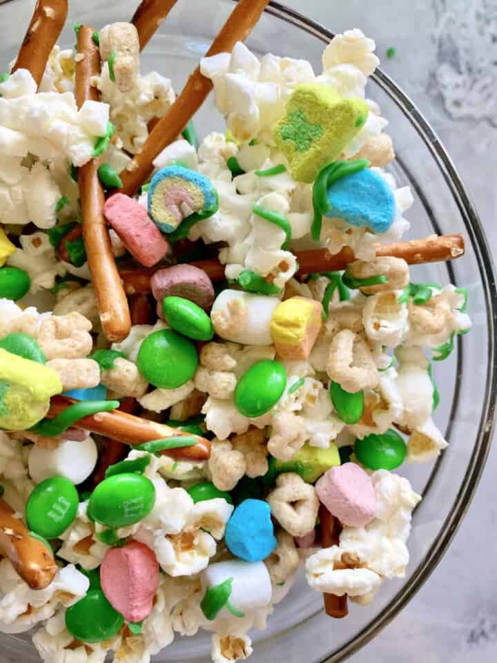 Close up of a glass bowl of popcorn, green M&M's, marshmallows, lucky charms, and pretzels.
