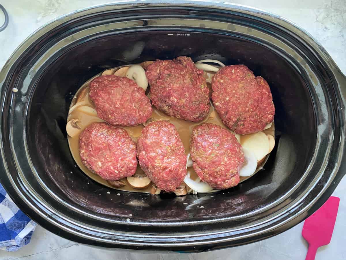 Top view of a black slow cooker with 6 ground beef paties resting on onions and broth.