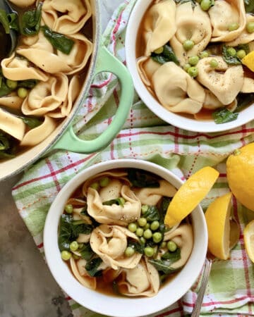 Top view of two white bowls and a green pot all filled with tortellini soup.