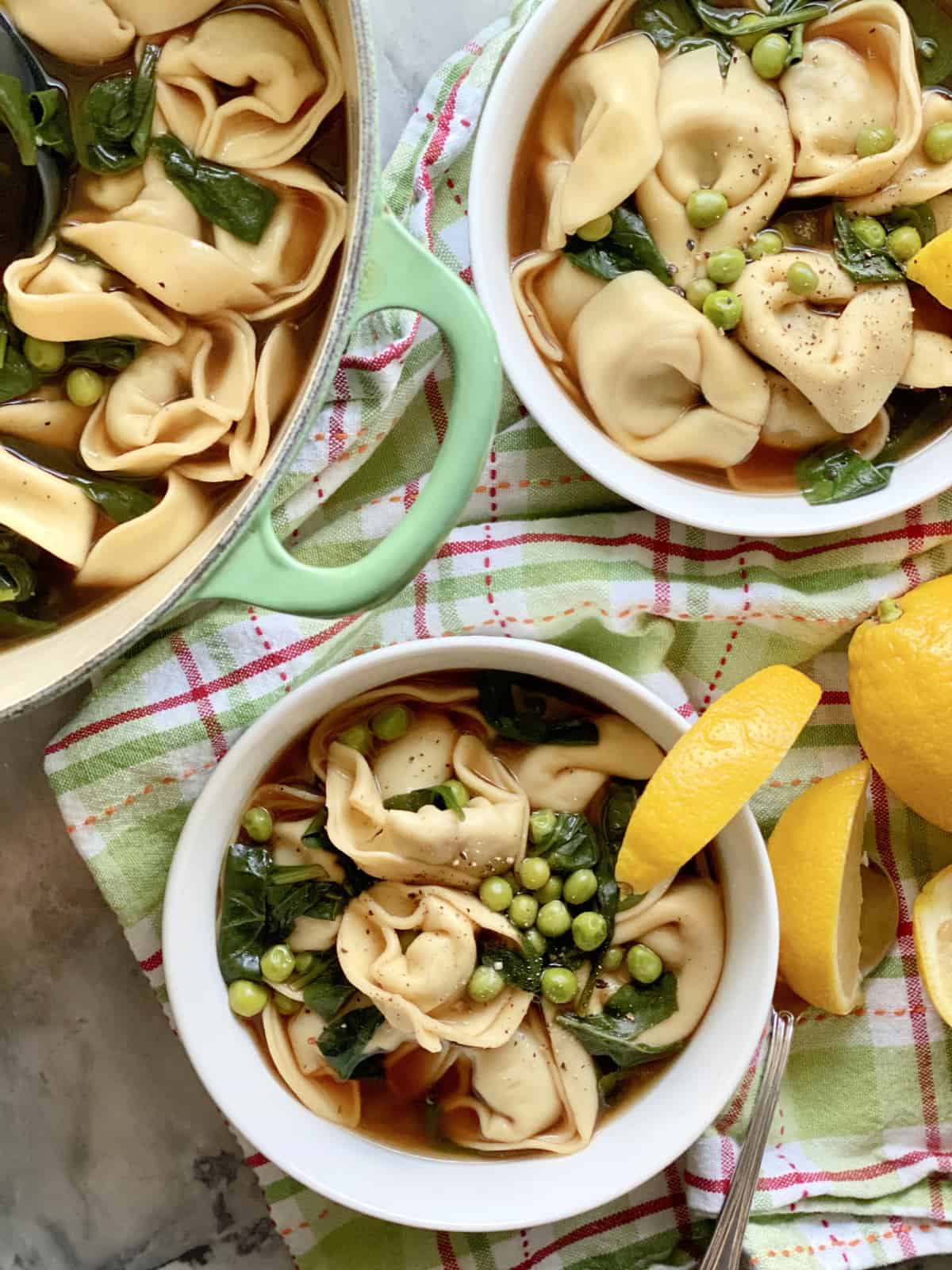 Top view of two white bowls and a green pot all filled with tortellini soup.