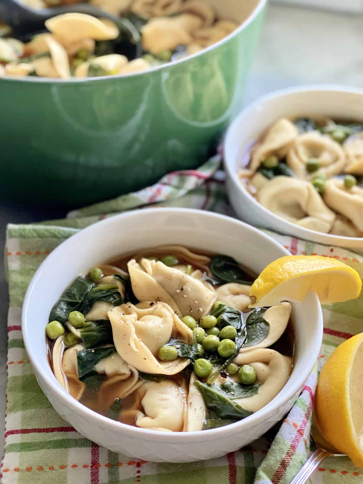 Bowl of tortellini soup with a lemon on the rim with a bowl and pot in the background.