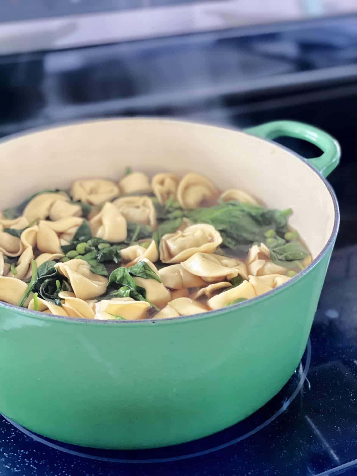 Green pot on a stove top with tortelini, spinach, and peas simmering with steam raised.