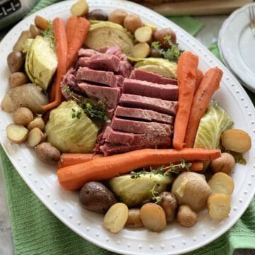 Top view of a white platter filled with meat and vegetables with plates and silverware on the side.