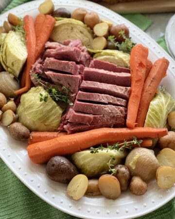 Top view of a white platter filled with meat and vegetables with plates and silverware on the side.