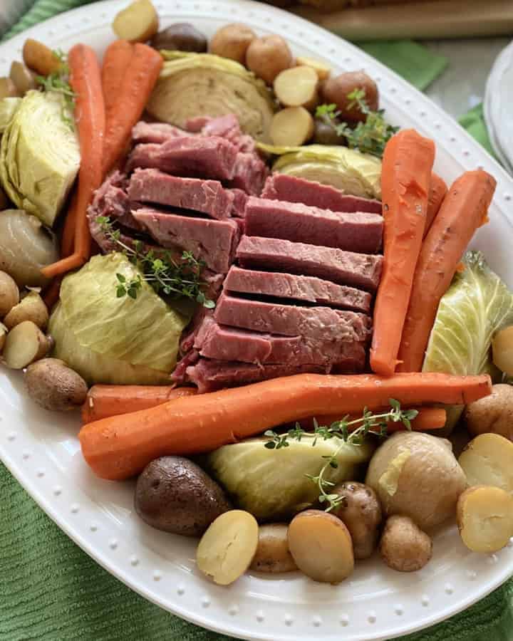 Top view of a white platter filled with meat and vegetables with plates and silverware on the side.