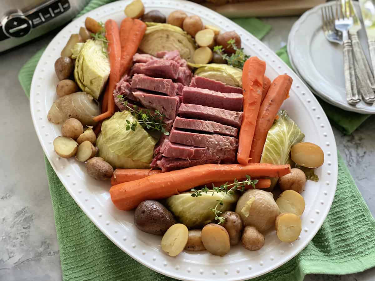 Top view of a white platter filled with meat and vegetables with plates and silverware on the side.