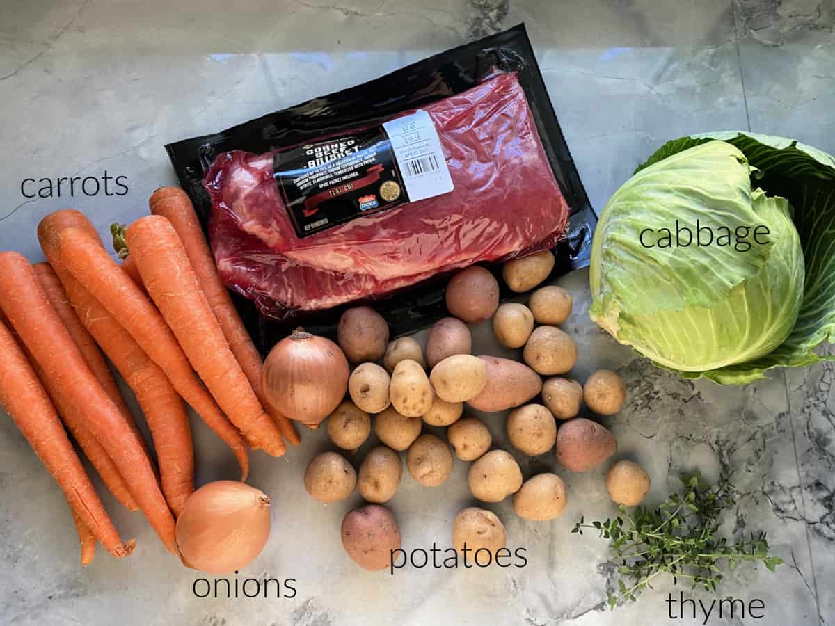 Ingredients on marble countertop: corned beef, carrots, potatoes, onions, cabbage, and thyme.