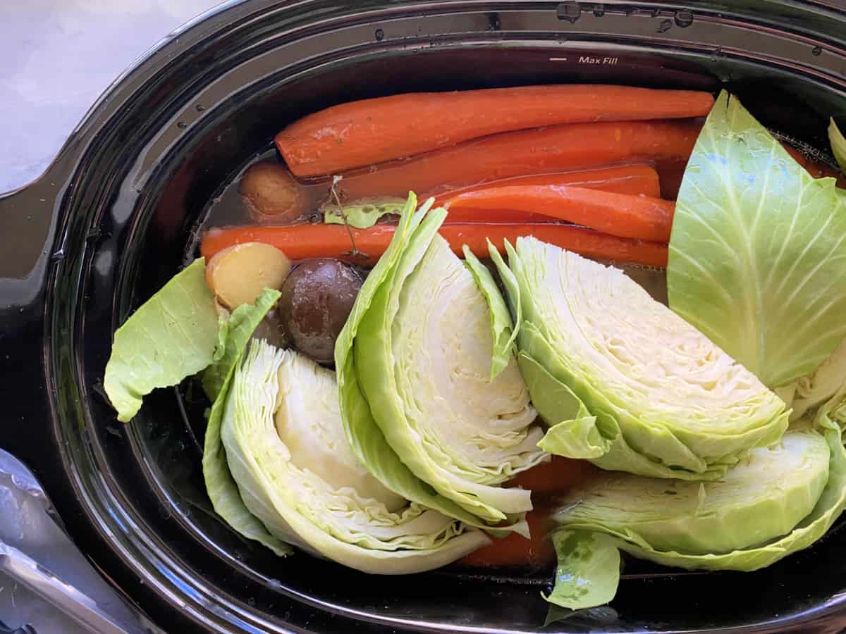 Top view of quartered cabbage, carrots, and potatoes in a black slow cooker.