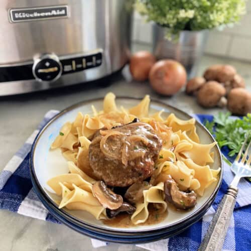 White plate filled with noodles and a hamburger steak with gravy and a stainless steel slow cooker in the background.