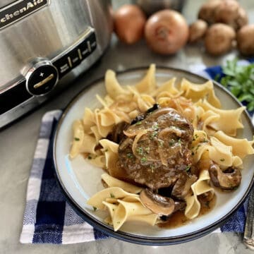 White plate filled with noodles, gravy, and beef steak on a blue checkered cloth napkin.