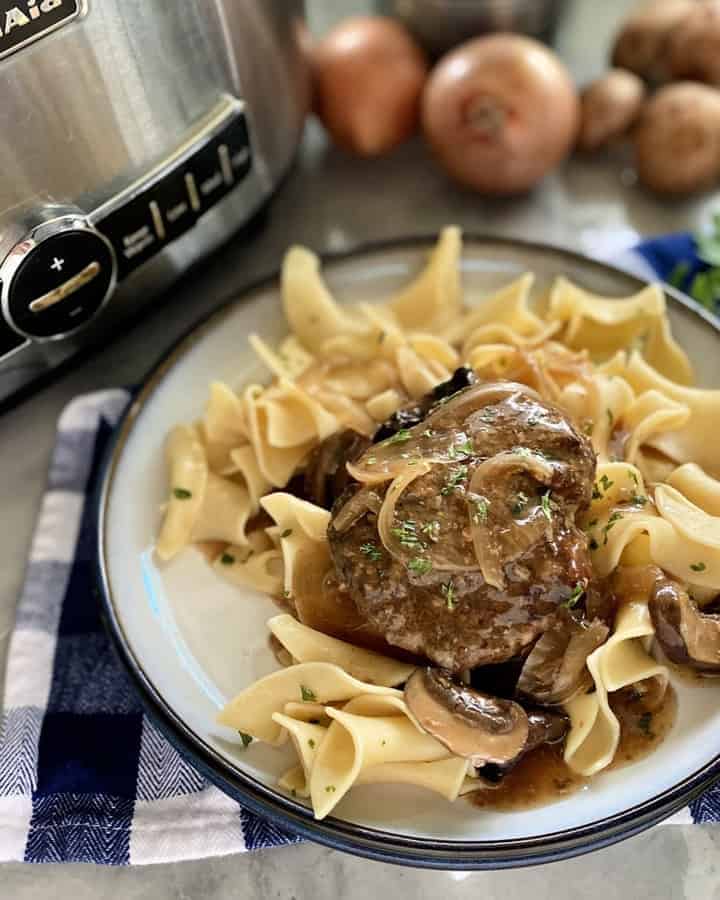 White plate filled with noodles, gravy, and beef steak on a blue checkered cloth napkin.