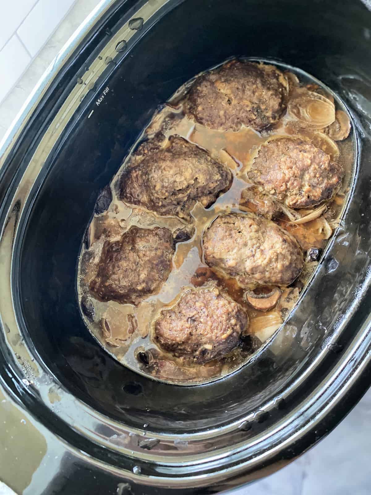 Top view of a black slow cooker filled with gravy and meat patties.