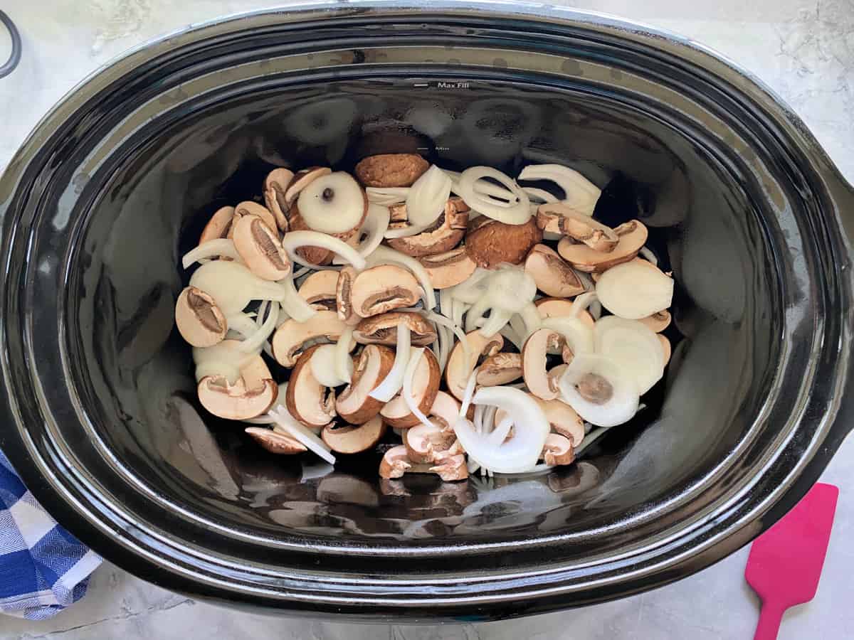 Top view of black crock pot with sliced mushrooms and onions.