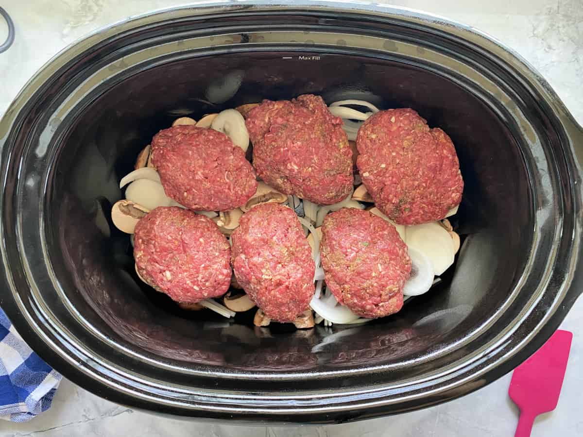 Top view of a slow cooker filled with onions and mushrooms with ground beef patties on top.