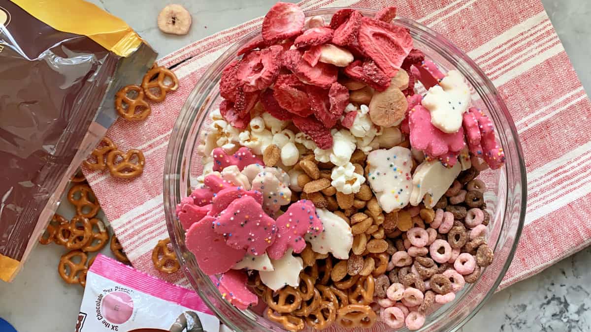 Top view of various snacks {pretzels, cheerios, popcorn, animal cookies) in a glass bowl.