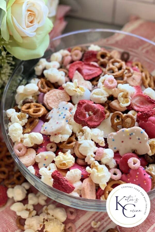 Glass bowl filled with pretzels, popcorn, animal crackers, and cheerios with logo on right corner.