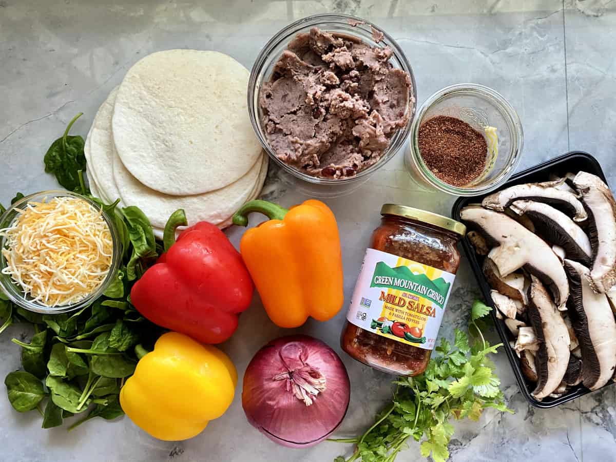 Ingredients on counter: tortillas, salsa, peppers, onions, mushrooms, fajita seasoning, cheese, spinach, black beans.