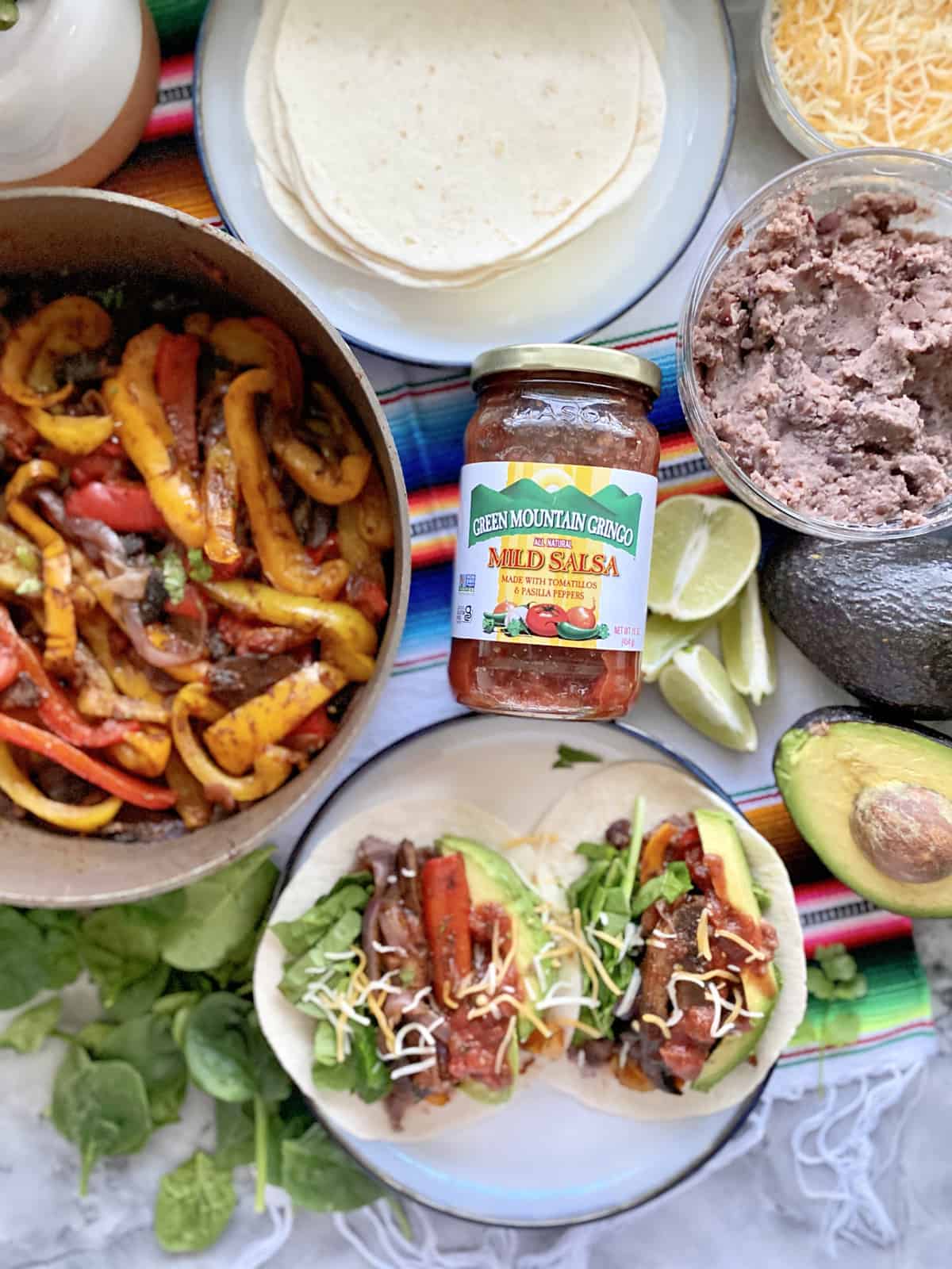 Glass jar of Green Mountain Gringo Salsa in focus with fajita accompiantments blurred in background.