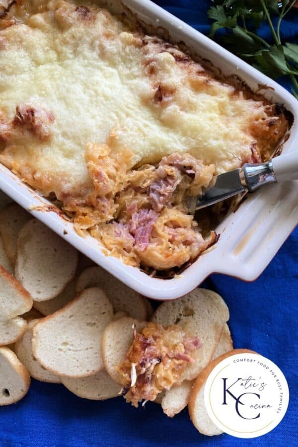 Top view of a white casserole dish filled with dip and a spreading knife with baguettes around the baking dish.