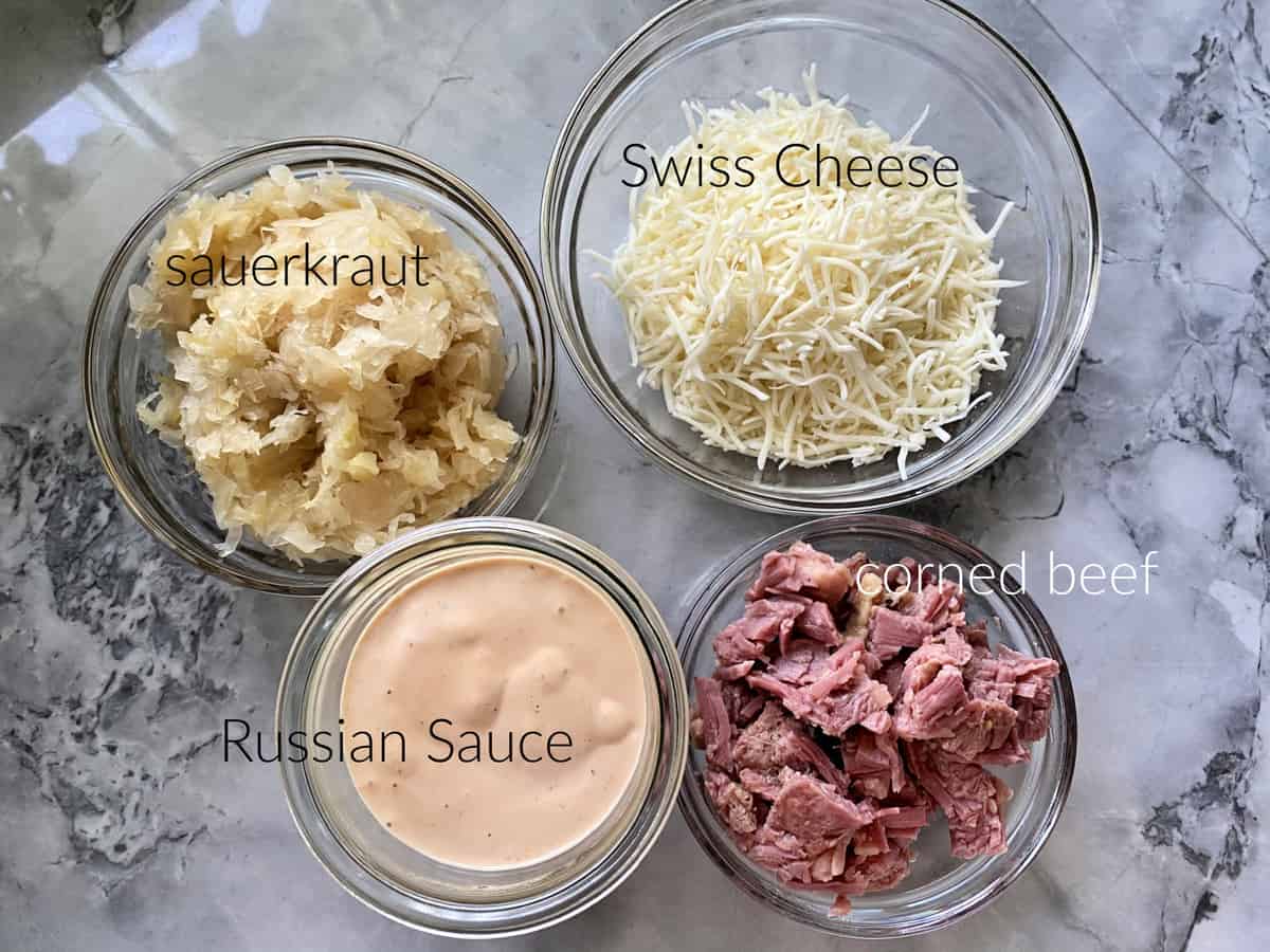 Ingredients on marble countertop: sauerkraut, swiss cheese, Russian sauce, and corned beef.