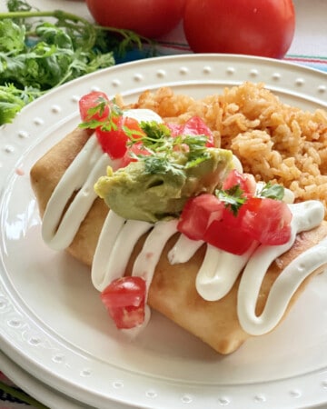 white plate with fried burrito topped with sour cream, gaucaumole, and diced tomatoes on top.