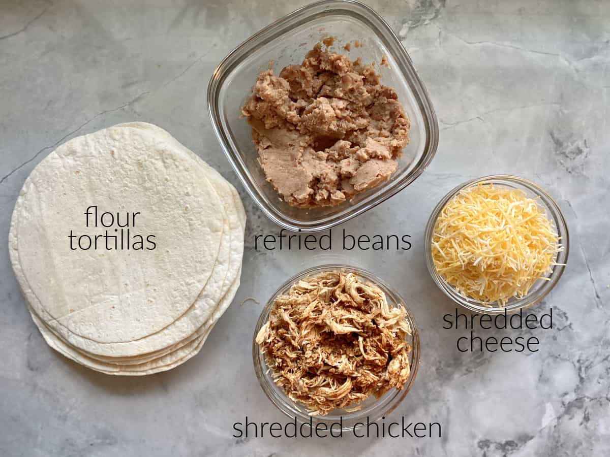 Ingredients on marble countertop: tortillas, refried beans, shredded chicken, shredded cheese.