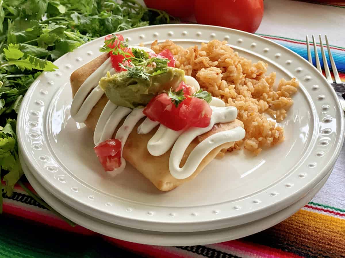 Two white plates stacked with a Chicken Chimichanga topped with sour cream, gucamole and tomatoes.