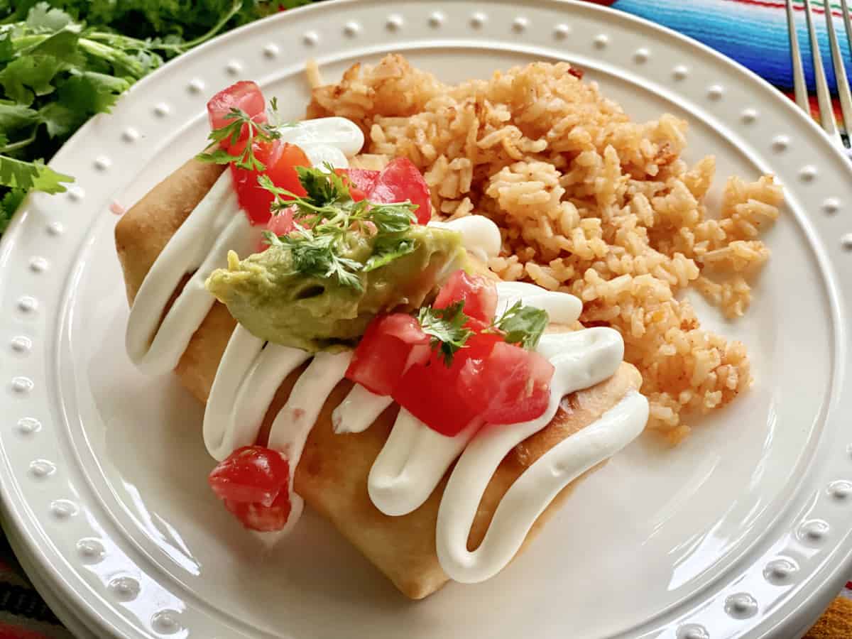 Fried burrito on a white plate topped with sour cream, tomatoes, and gucamole.