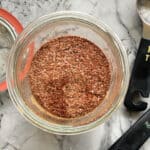 Top view of a glass filled with seasoning and measuring spoons laying on marble counter next to glass jar.