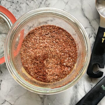 Top view of a glass filled with seasoning and measuring spoons laying on marble counter next to glass jar.