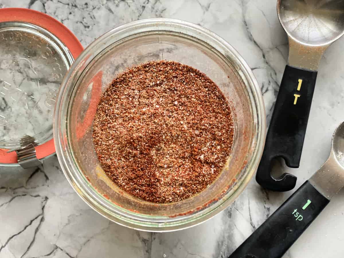 Top view of a glass filled with seasoning and measuring spoons laying on marble counter next to glass jar.