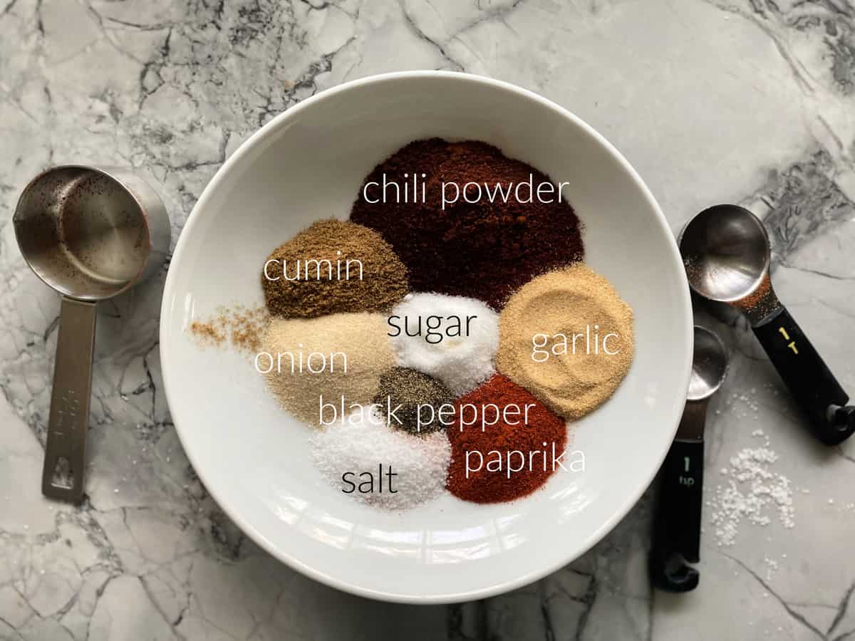 Ingredients in shallow bowl: chili powder, cumin, sugar, onion, garlic, black pepper, salt, and paprika.