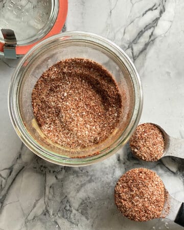 Top view of a glass jar filled with seasoning and two measuring spoons on marble countertop with seasoning.