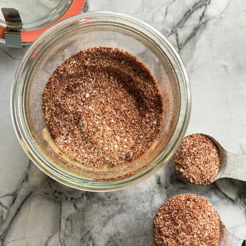 Top view of a glass jar filled with seasoning and two measuring spoons on marble countertop with seasoning.