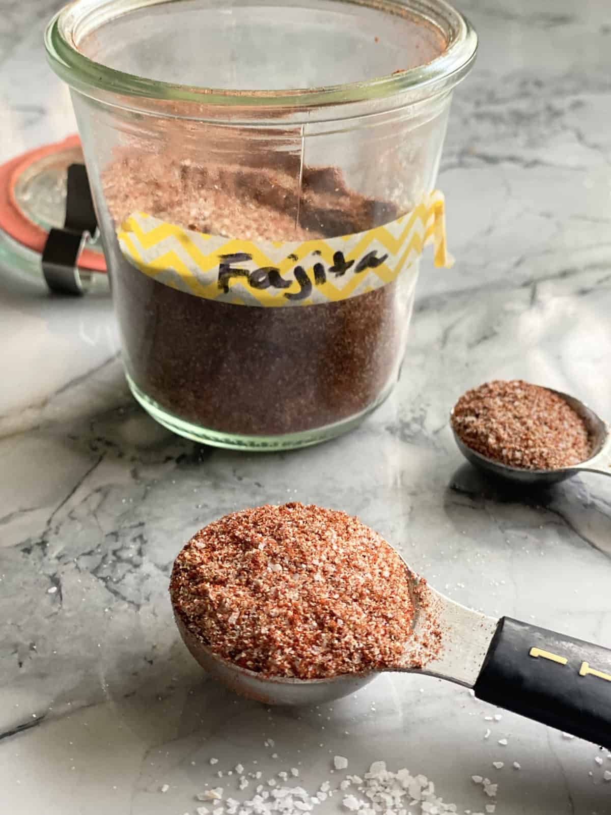 Glass jar with yellow tape label "fajita" with tablespoon filled with seasoning on countertop.
