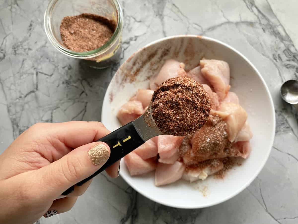 Female hand with glitter nail polish holding a tablespoon full of fajita seasoning over raw diced chicken.