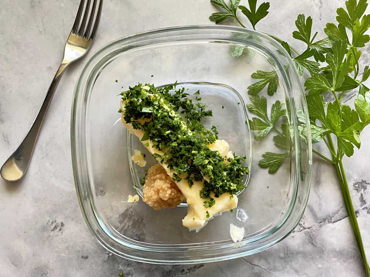 Square glass bowl filled with a stick of butter, minced parsley, and garlic.