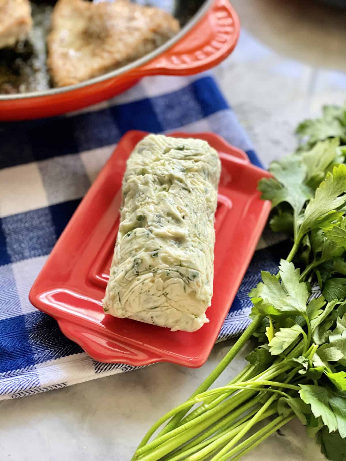 Red dish filled with a log of Garlic Herb Butter sitting on a blue and white checkered cloth.