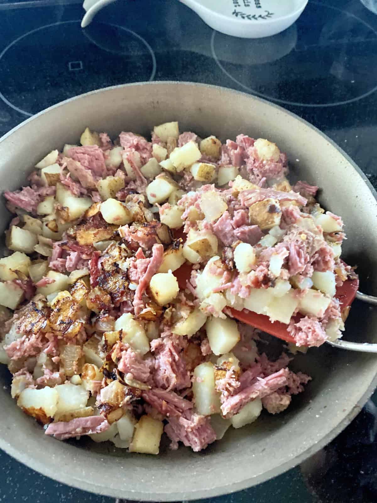 Skillet filled with Corned Beef Hash with red spatula picking up the hash to flip.