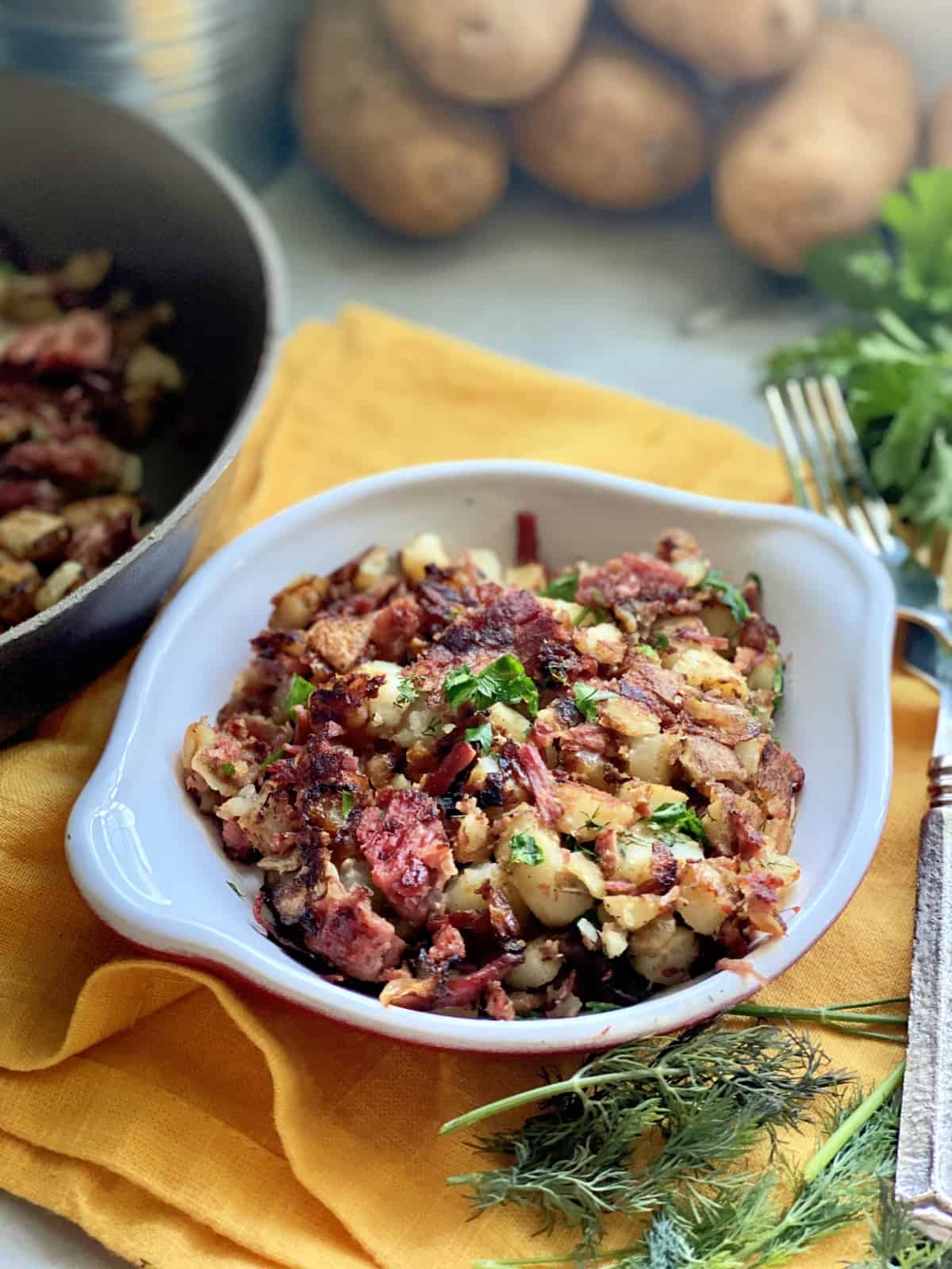 White shallow dish filled with corned beef hash with potatoes and skillet in background.